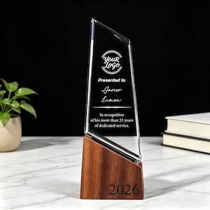 Op de afbeelding: Een heldere glazen award met een houten voet. De award heeft tekst met "Your Logo", "Presented to Javier Limon" en "In recognition of his more than 25 years of dedicated service." Het nummer "2026" staat op de voet.