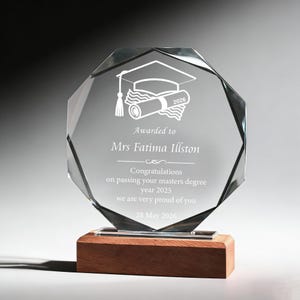 May include: A clear, faceted glass award on a wooden base. The award has an etched graduation cap and scroll design, with the text "Awarded to Mrs Fatima Illston" and a congratulatory message. The date "28 May 2026" is also included.