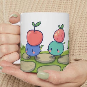 Könnte beinhalten: Weiße Keramik-Tasse mit einer Cartoon-Illustration von zwei Figuren, eine mit einem Apfel und einer Blaubeere, die andere mit einer Erdbeere und einer türkisfarbenen Figur. Die Figuren gehen auf Trittsteinen.