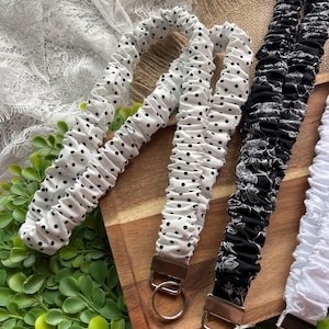 May include: A collection of fabric lanyards featuring gathered, elasticized designs. One lanyard is white with black polka dots, another is black with a white floral pattern, and a third is solid white. Each includes a metal clasp and ring.
