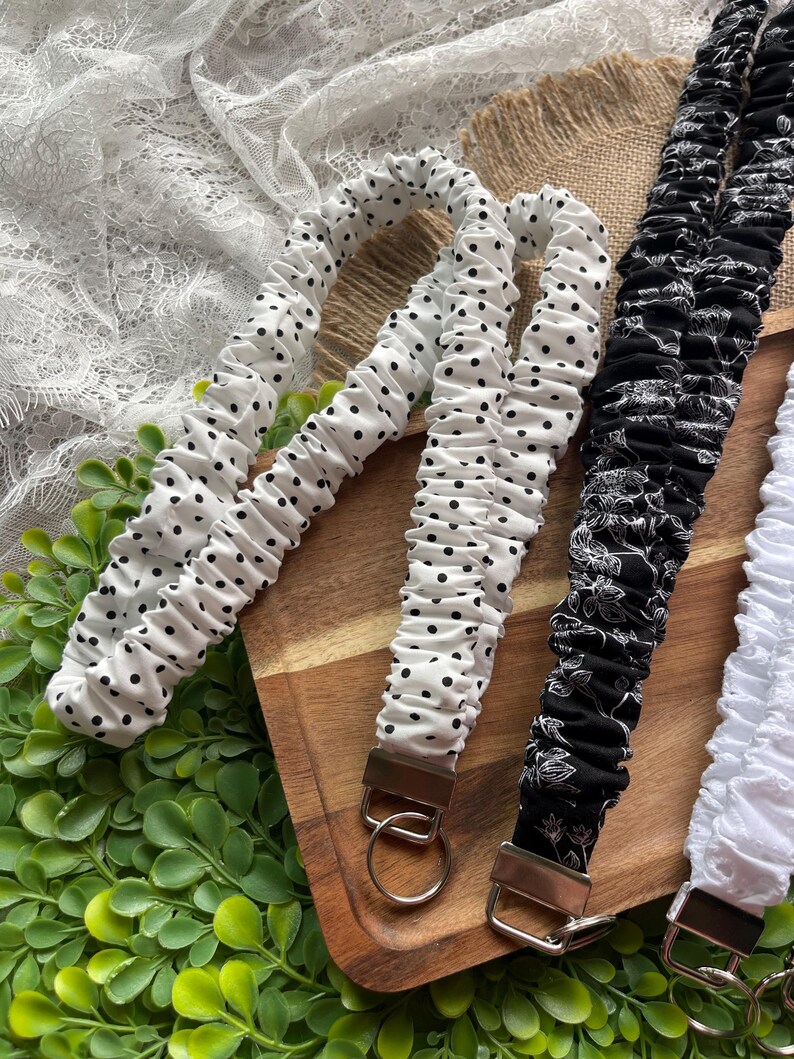 May include: A collection of fabric lanyards featuring gathered, elasticized designs. One lanyard is white with black polka dots, another is black with a white floral pattern, and a third is solid white. Each includes a metal clasp and ring.