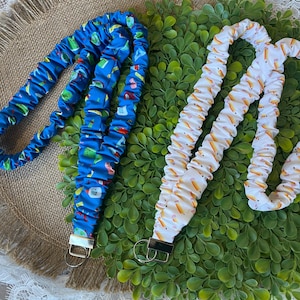 May include: Two fabric lanyards. One is blue with a pattern of various objects. The other is white with a pattern of yellow and orange pencils. Each lanyard has a metal clasp and a metal key ring.