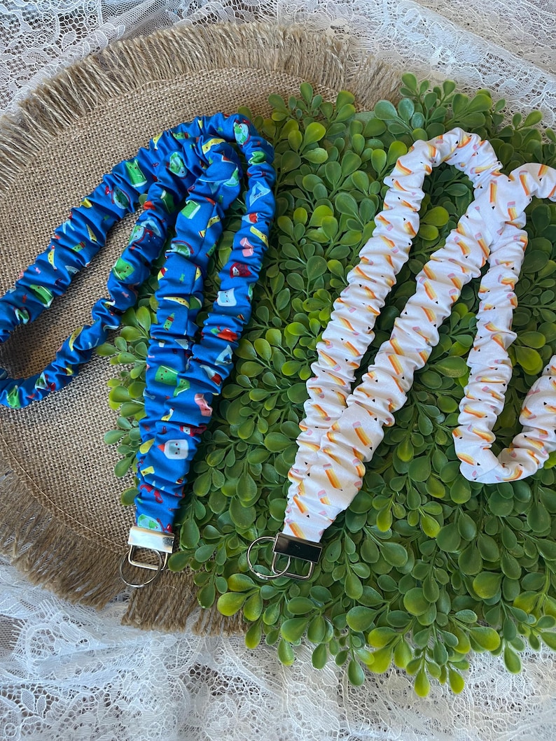 May include: Two fabric lanyards. One is blue with a pattern of various objects. The other is white with a pattern of yellow and orange pencils. Each lanyard has a metal clasp and a metal key ring.