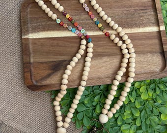 May include: Two wooden bead lanyards with colorful letter beads spelling out "MRS" and "FIRST GRADE". The lanyards have bronze-colored swivel clasps and large wooden beads. The lanyards are displayed on a wooden tray with greenery.