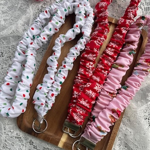 May include: A collection of fabric lanyards in various festive patterns, including red with white snowflakes, white with red and green polka dots, and pink with gingerbread men. Each lanyard has a silver-toned metal clasp.