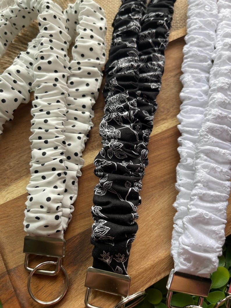 May include: Three fabric lanyards with silver-tone metal hardware and key rings. One is white with black polka dots, one is black with a white floral line-art pattern, and one is solid white with a textured, eyelet-style fabric finish.