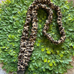 May include: A leopard print lanyard with a metal clasp. The lanyard is brown and tan with dark brown spots. The lanyard is lying on a green leafy wreath with a burlap border.
