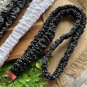May include: A collection of fabric lanyards featuring gathered, elasticized designs. The set includes black lanyards with white polka dots, a white textured lanyard, and a black lanyard with a white floral pattern, each with a silver metal clasp.