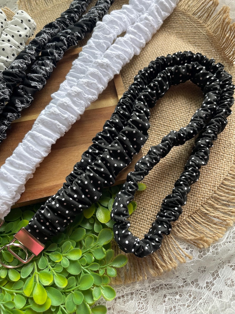 May include: A collection of fabric lanyards featuring gathered, elasticized designs. The set includes black lanyards with white polka dots, a white textured lanyard, and a black lanyard with a white floral pattern, each with a silver metal clasp.