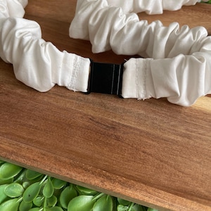 May include: A white fabric belt featuring a gathered, elasticized design that creates a ruffled texture throughout the length of the band. The belt is secured in the center with a rectangular black plastic buckle clasp that connects the two ends.