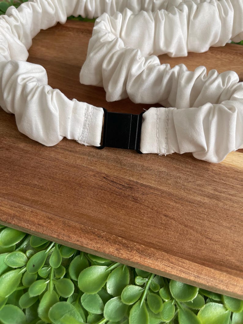 May include: A white fabric belt featuring a gathered, elasticized design that creates a ruffled texture throughout the length of the band. The belt is secured in the center with a rectangular black plastic buckle clasp that connects the two ends.
