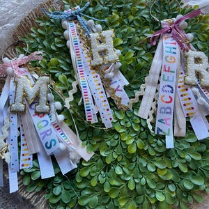 May include: Three keychains with the letters "M", "H", and "R". Each keychain has a letter with a gold trim, attached to colorful ribbons and white pom-poms. The ribbons have text and patterns. The keychains are on a green wreath.