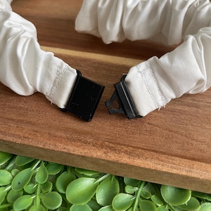 May include: A white fabric headband featuring a gathered, elasticized design with a black plastic side-release buckle closure. The white material is bunched along the length of the band, creating a textured appearance that connects at the black clasp.