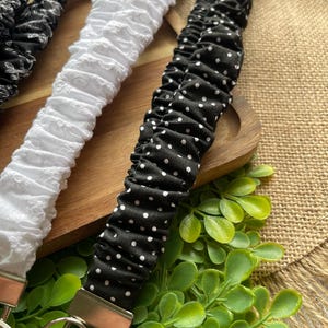 May include: Three fabric wristlet keychains featuring gathered, elasticized designs. One is black with a white floral pattern, one is solid white with a textured eyelet finish, and one is black with a small white polka dot pattern, each with a metal ring.