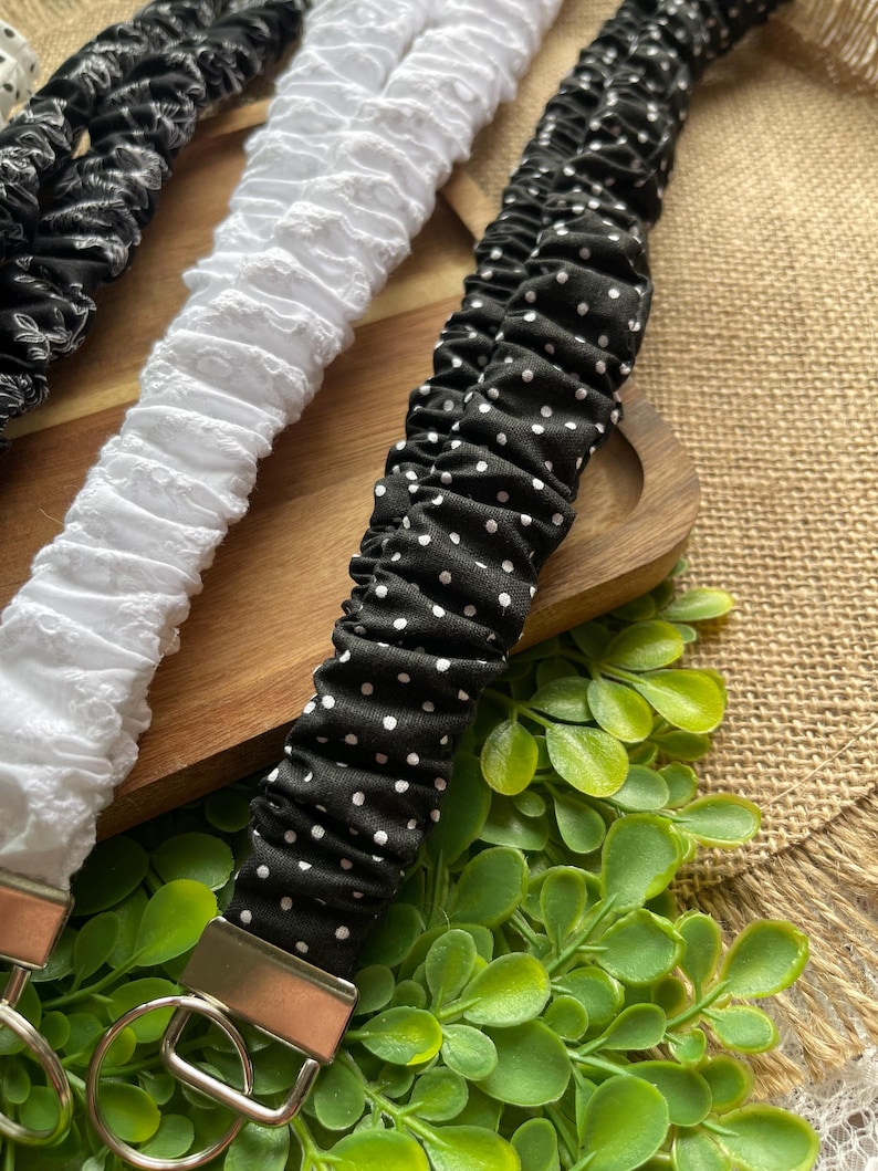 May include: Three fabric wristlet keychains featuring gathered, elasticized designs. One is black with a white floral pattern, one is solid white with a textured eyelet finish, and one is black with a small white polka dot pattern, each with a metal ring.