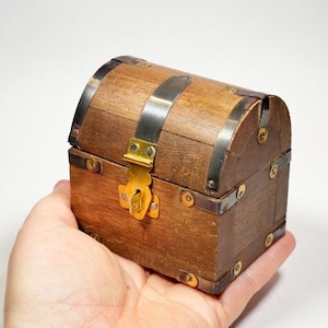May include: A small, wooden treasure chest with a curved top and metal bands. The chest has a brass clasp and hinges. The wood is a warm brown color, and the metal accents are silver. The chest is held in a hand.