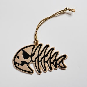 May include: A gold-colored fish skeleton ornament with black detailing and a gold string for hanging. The fish has a black eye patch, giving it a playful, pirate-like appearance. The ornament is likely for decorative purposes.