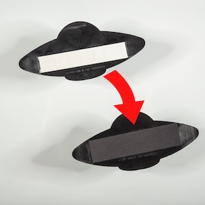 May include: Two black, oval-shaped objects with a central protrusion. The top object features a white adhesive strip on its flat side. A red arrow points downward to the bottom object, which displays a black magnetic strip on its flat side.