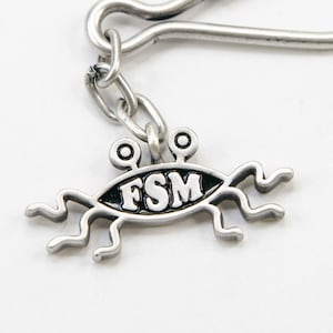 May include: A silver-colored keychain charm featuring a stylized depiction of the Flying Spaghetti Monster. The charm has a black oval body with the letters "FSM" in white. It has two eye-like protrusions and several noodle-like appendages.