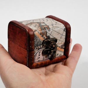 May include: A small, wooden treasure chest with a map design on the lid. The chest is rectangular with dark brown sides and a metal clasp. The map design features shades of brown and beige, giving it a vintage look.