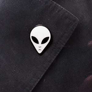May include: A white alien face pin with black eyes and a black outline is pinned on a black fabric. The pin is a unique accessory, perfect for adding a touch of extraterrestrial flair to clothing or bags.