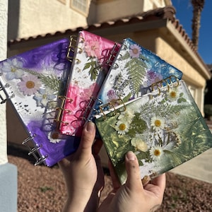 May include: Handmade resin notebooks with dried flowers embedded in the covers. The notebooks are in various colours: purple, pink, blue, and green, with gold spiral binding. Each notebook features a unique floral arrangement, perfect for journaling or note-taking.