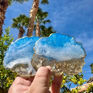 Puede incluir: Dos posavasos de resina con un diseño de temática playera. Los posavasos presentan un efecto de olas azules y blancas, con una base de arena y un borde dorado. Hay palmeras y un cielo azul en el fondo.