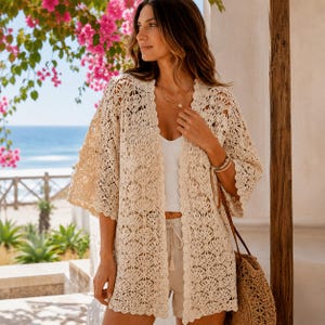 Peut inclure: Un gilet beige ouvert sur le devant, doté d'un motif en crochet complexe avec des bords festonnés et des manches larges trois-quarts. Le vêtement est porté sur un débardeur blanc et un short clair, accompagné d'un sac à bandoulière en paille tressée.