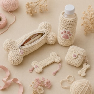 Peut inclure: Un ensemble d'accessoires en crochet couleur crème comprenant une trousse en forme d'os avec fermeture éclair, une housse de bouteille avec une patte rose, un marque-page en forme d'os à pompons roses et un étui à écouteurs avec un petit cœur rose.