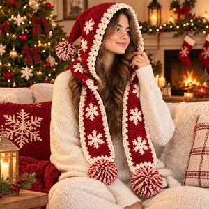 Peut inclure: Une écharpe à capuche rouge foncé avec une bordure en crochet crème et des motifs de flocons de neige blancs. L'écharpe comprend une capuche pointue et deux longues extrémités, chacune terminée par un gros pompon rond rouge et crème.