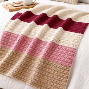 Peut inclure: Une couverture crochetée à rayures faite à la main, dans les tons beige, bordeaux, rose et beige. La couverture est pliée sur un lit blanc, avec un oreiller assorti en arrière-plan. La texture de la couverture est visible, mettant en valeur les points de crochet.