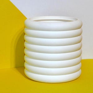 May include: A white, cylindrical ceramic planter with a ribbed design. The planter sits on a yellow surface, with a white and yellow background. The planter is a decorative item for indoor or outdoor use.