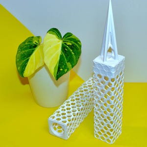 Puede incluir: Soportes de plantas geométricos blancos con diseño de panal, junto a una planta en maceta con hojas verdes y amarillas. El fondo es una división de amarillo y blanco. Los soportes están diseñados para plantas.