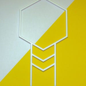 May include: A white, geometric plant support with a hexagonal top and a series of downward-pointing chevrons. The support has two pointed stakes at the bottom. The background is divided diagonally, with white and yellow sections.