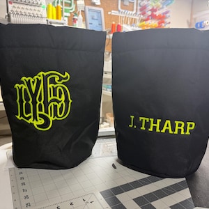 May include: Two black drawstring bags featuring lime green embroidery. The left bag displays a stylized monogram, while the right bag features the text J. THARP in a sans-serif font. Both bags have black drawstrings and attached product tags.