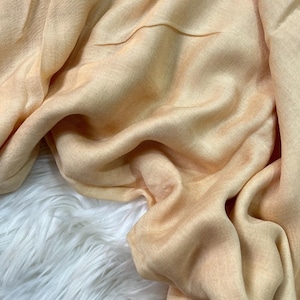May include: A close-up of a peach-colored scarf with a soft, flowing texture. The fabric is draped over a white, fluffy surface. The scarf has subtle variations in color, creating a gentle gradient effect.