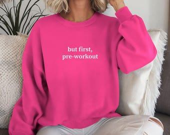 May include: A vibrant pink sweatshirt with the text "but first, pre-workout" in white. The long-sleeved top is made of a soft material and is perfect for workouts or casual wear.