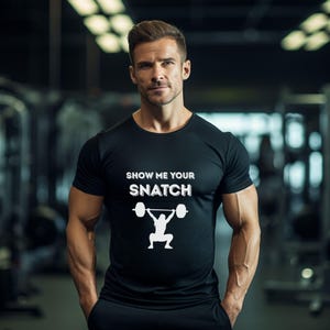 May include: Black t-shirt with the text "SHOW ME YOUR SNATCH" in white, along with a weightlifting graphic. The shirt is worn by a person in a gym setting.