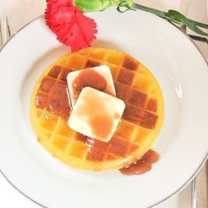 May include: A white plate with a waffle topped with butter and brown sauce. A red carnation is in the background.
