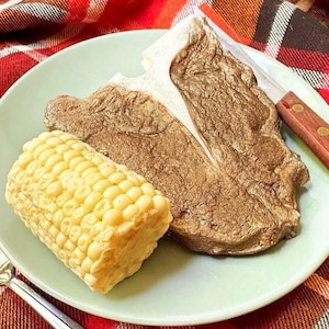 May include: A green plate with a cooked T-bone steak and a corn on the cob. The plate is on a red and white plaid tablecloth.