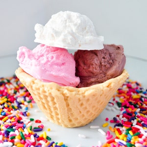 May include: A waffle cone filled with three scoops of ice cream: pink, white, and chocolate. The ice cream is surrounded by colorful sprinkles.