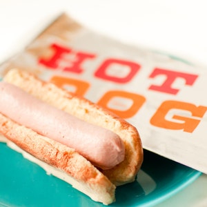 May include: A hot dog in a bun on a teal plate, with a paper bag in the background. The hot dog is light pink, and the bun is toasted. The paper bag has the words "HOT DOG" printed in red and orange.