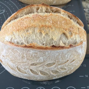May include: A freshly baked loaf of sourdough bread with a golden-brown crust. The bread has a decorative leaf pattern carved into the bottom half. The top crust is cracked and textured, indicating a crispy exterior. The bread is on a dark gray surface.