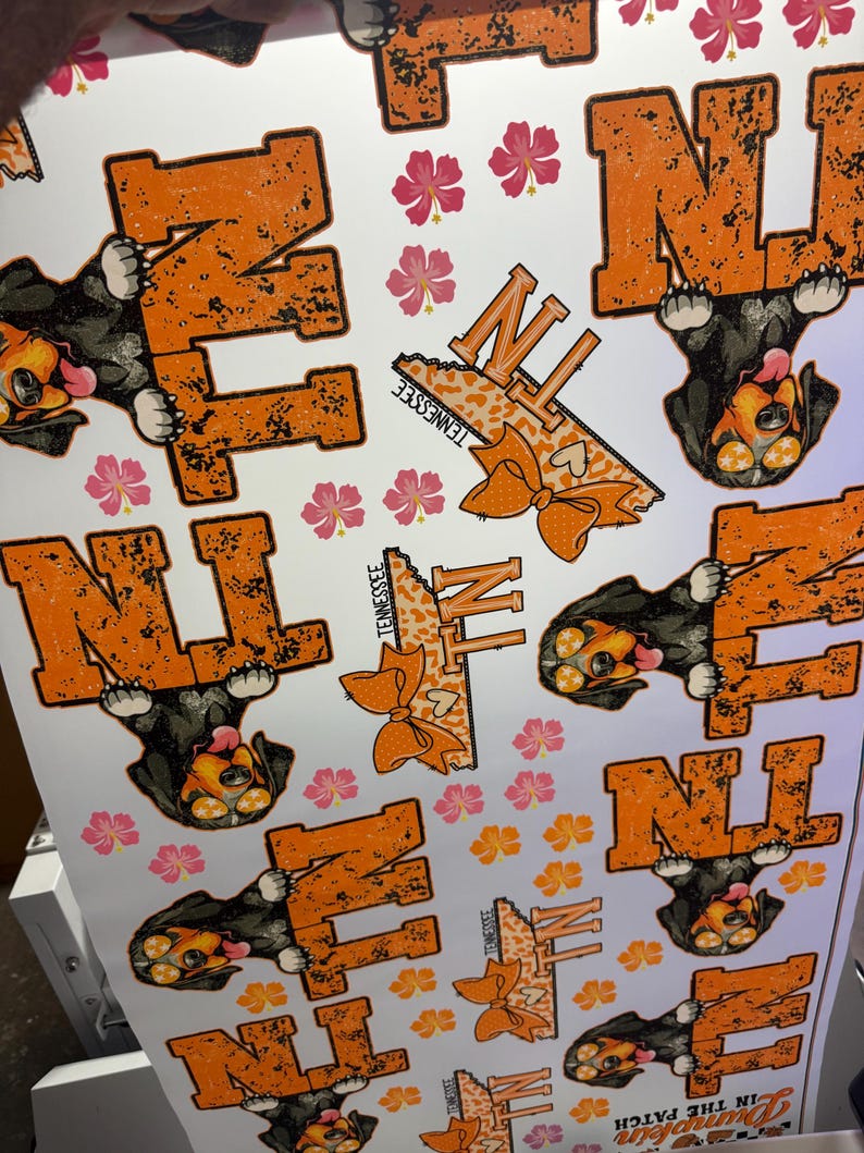 K&ouml;nnte beinhalten: Wei&szlig;es bedrucktes Blatt mit orangefarbenen "TN"-Buchstaben mit einem Used-Look, einem Cartoon-Hund und rosa und orangefarbenen floralen Akzenten. Das Wort "Tennessee" ist ebenfalls aufgedruckt.