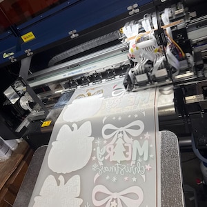 K&ouml;nnte beinhalten: Ein Gro&szlig;formatdrucker in Betrieb, der weihnachtliche Designs auf eine Materialrolle druckt. Die Designs umfassen Schleifen, Weihnachtsb&auml;ume und den Text "Merry Christmas" in einer festlichen Schriftart. Der Drucker ist blau und schwarz.