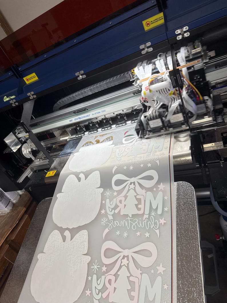 K&ouml;nnte beinhalten: Ein Gro&szlig;formatdrucker in Betrieb, der weihnachtliche Designs auf eine Materialrolle druckt. Die Designs umfassen Schleifen, Weihnachtsb&auml;ume und den Text "Merry Christmas" in einer festlichen Schriftart. Der Drucker ist blau und schwarz.