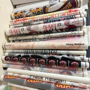 K&ouml;nnte beinhalten: Ein Stapel gerollter Drucke, jeweils in transparentes Papier gewickelt. Die Drucke zeigen verschiedene Designs, darunter Texte wie "LIKE", "ROS" und Namen wie "Mari Santiago" und "Avery Barrows". Die Farbpalette umfasst Rot, Schwarz, Wei&szlig; und Grau.