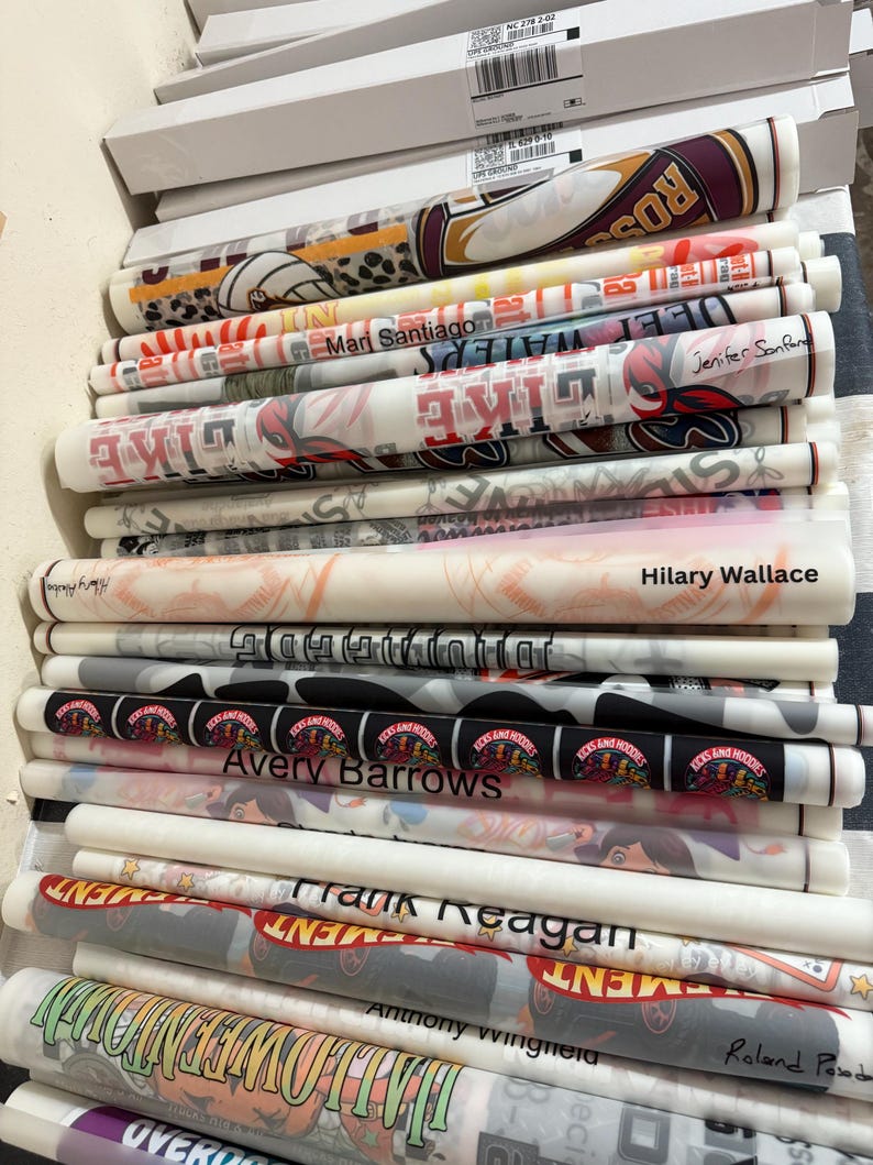 K&ouml;nnte beinhalten: Ein Stapel gerollter Drucke, jeweils in transparentes Papier gewickelt. Die Drucke zeigen verschiedene Designs, darunter Texte wie "LIKE", "ROS" und Namen wie "Mari Santiago" und "Avery Barrows". Die Farbpalette umfasst Rot, Schwarz, Wei&szlig; und Grau.