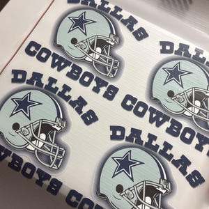 K&ouml;nnte beinhalten: Bogen mit Aufklebern von Dallas Cowboys-Helmen. Die Aufkleber zeigen einen hellblauen Helm mit einem marineblauen Stern und den Worten "DALLAS COWBOYS" in Marineblau. Auf wei&szlig;em Hintergrund.