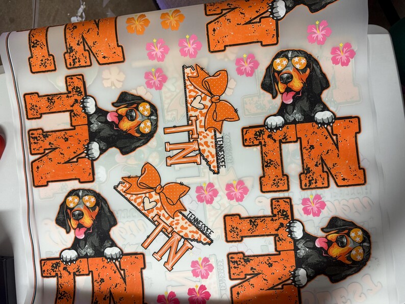 K&ouml;nnte beinhalten: Ein Bogen mit Aufklebern mit den Buchstaben "TN" in Orange mit einem Used-Look und Bildern eines Hundes mit Sonnenbrille. Die Aufkleber enthalten auch den Bundesstaat Tennessee mit einer orangefarbenen Schleife und Hibiskusbl&uuml;ten.